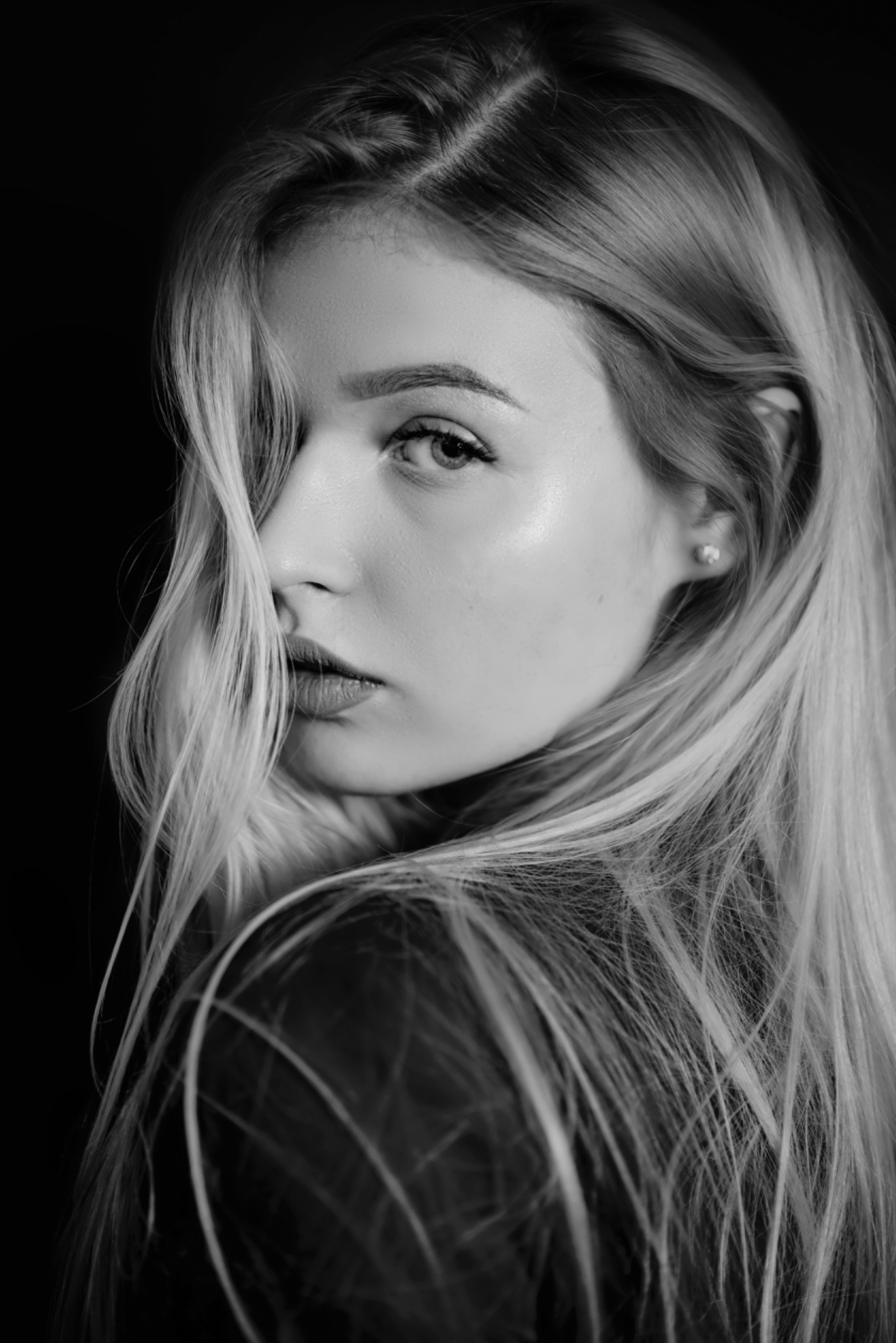 Portret czarno-biały kobiety z długimi, jasnymi włosami, spoglądającej przez ramię, z widoczną delikatną biżuterią w uchu i subtelnym makijażem.
