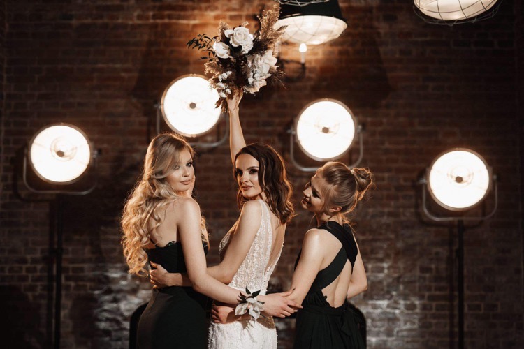 Panna młoda w białej sukni z bukietem kwiatów, otoczona dwiema druhnami w czarnych sukniach, pozuje na tle ceglanej ściany oświetlonej studyjnymi lampami.