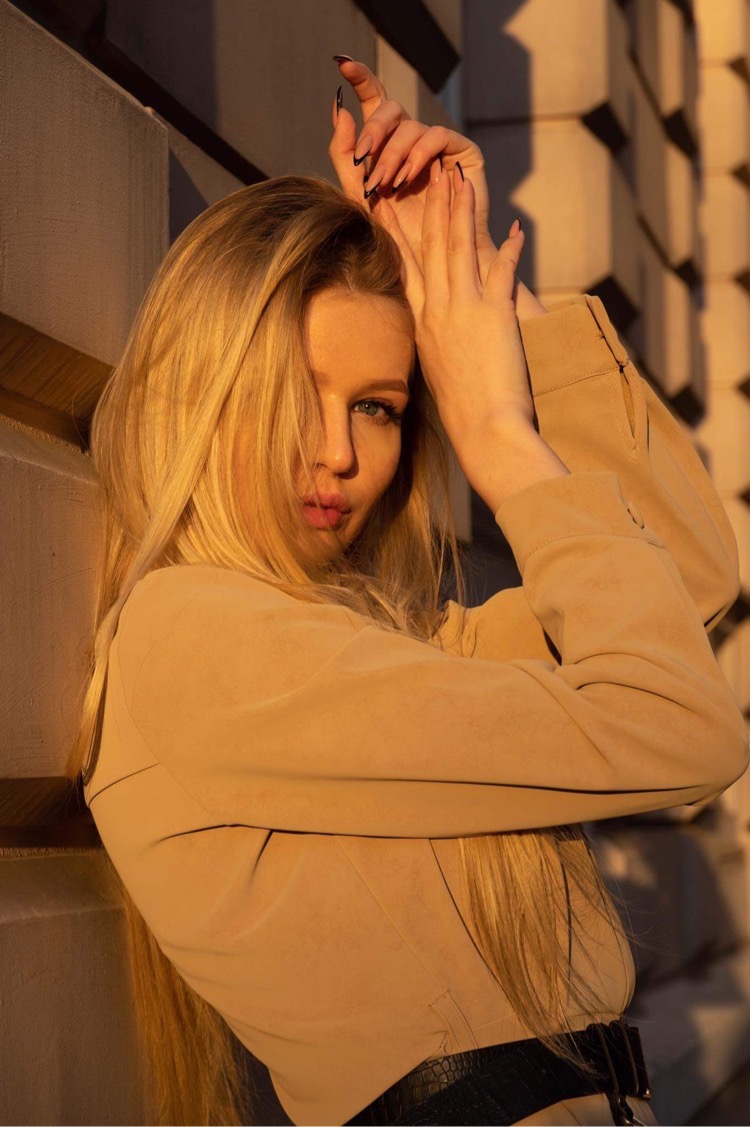 Portret kobiety z blond włosami, w beżowej marynarce i czarnym pasku, opierającej się o ścianę w ciepłym, zachodzącym słońcu, z dłońmi uniesionymi nad głową.