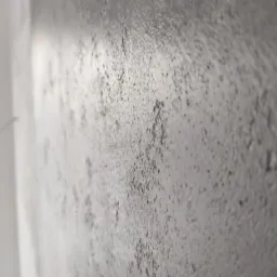 Szara, chropowata ściana z efektem betonu, sfotografowana pod kątem, z widocznym fragmentem białej listwy przypodłogowej.