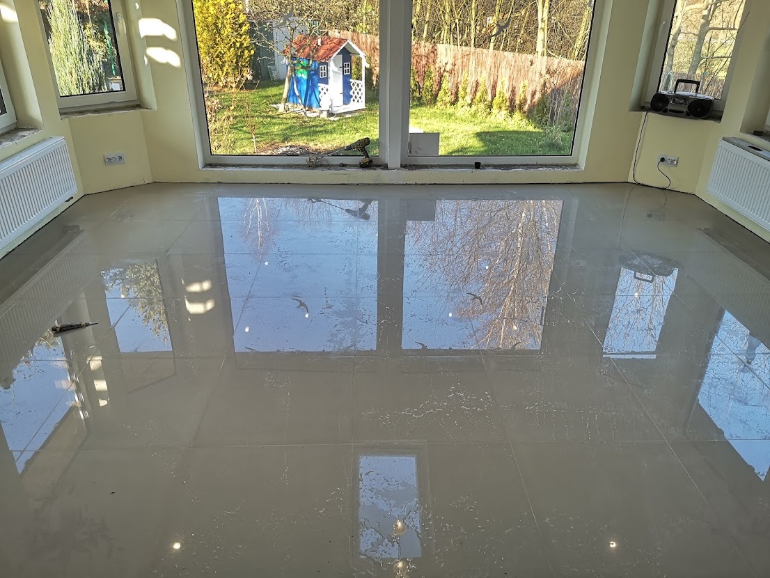 Błyszcząca, jasna podłoga z płytek ceramicznych w nowo wykończonym pomieszczeniu z dużymi oknami i widokiem na ogród; widoczne narzędzia i ślady po fugowaniu.