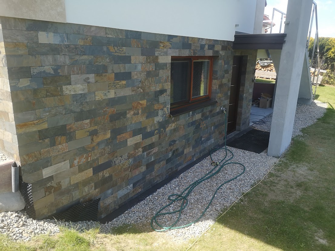 Elewacja budynku wykończona płytkami imitującymi kamień naturalny w odcieniach szarości, brązu i złota, widoczne okno z brązową ramą, zielony wąż ogrodowy i fragment trawnika.