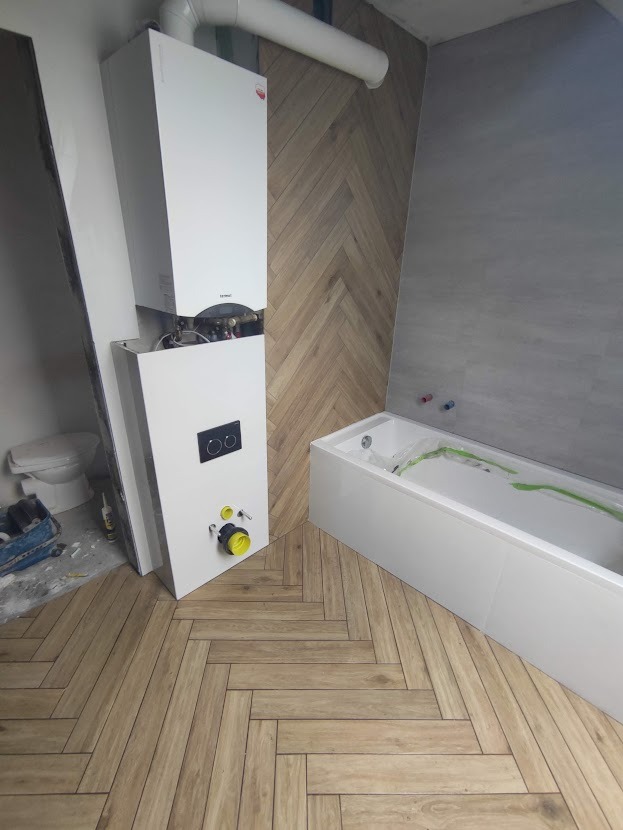 Niedokończona łazienka z zamontowanym piecem dwufunkcyjnym, białą wanną zabezpieczoną folią i drewnopodobną podłogą ułożoną w jodełkę.