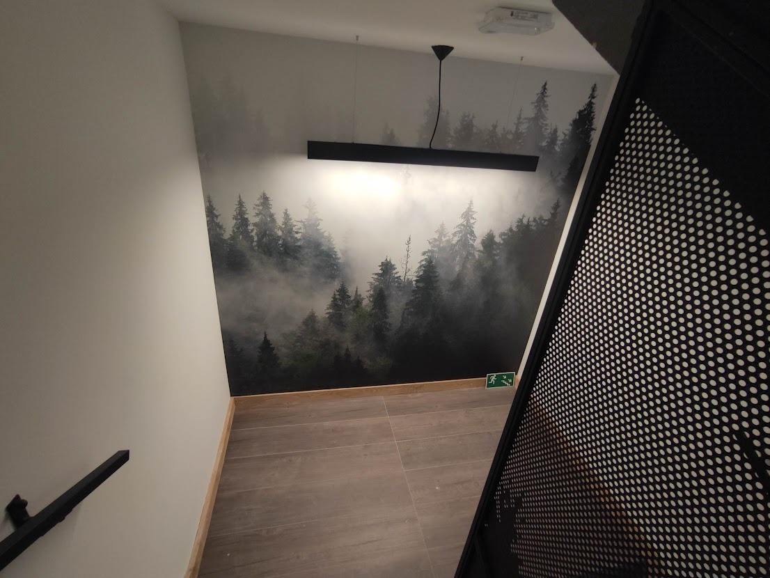 Klatka schodowa z fototapetą przedstawiającą las we mgle, oświetlona nowoczesną, podłużną lampą wiszącą i z metalową ażurową balustradą.
