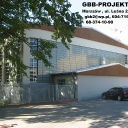 Dwukondygnacyjny budynek z przeszkloną fasadą i brązowym dachem, widoczny z zewnątrz, z zaparkowanymi samochodami i metalowym ogrodzeniem, częściowo zasłonięty przez drzewo.