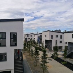 Tomasz Kiszko - Osiedle domów jednorodzinnych w zabudowie szeregowej z białymi elewacjami i czarnymi ramami okiennymi, widoczne schody zewnętrzne i młode drzewka iglaste wzdłuż ścieżki.