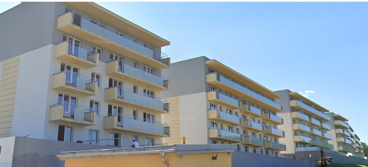 Nowoczesne osiedle budynków wielorodzinnych z balkonami o szklanych balustradach, widok z zewnątrz w słoneczny dzień, jeden z pracowników siedzi na dachu niskiego budynku.