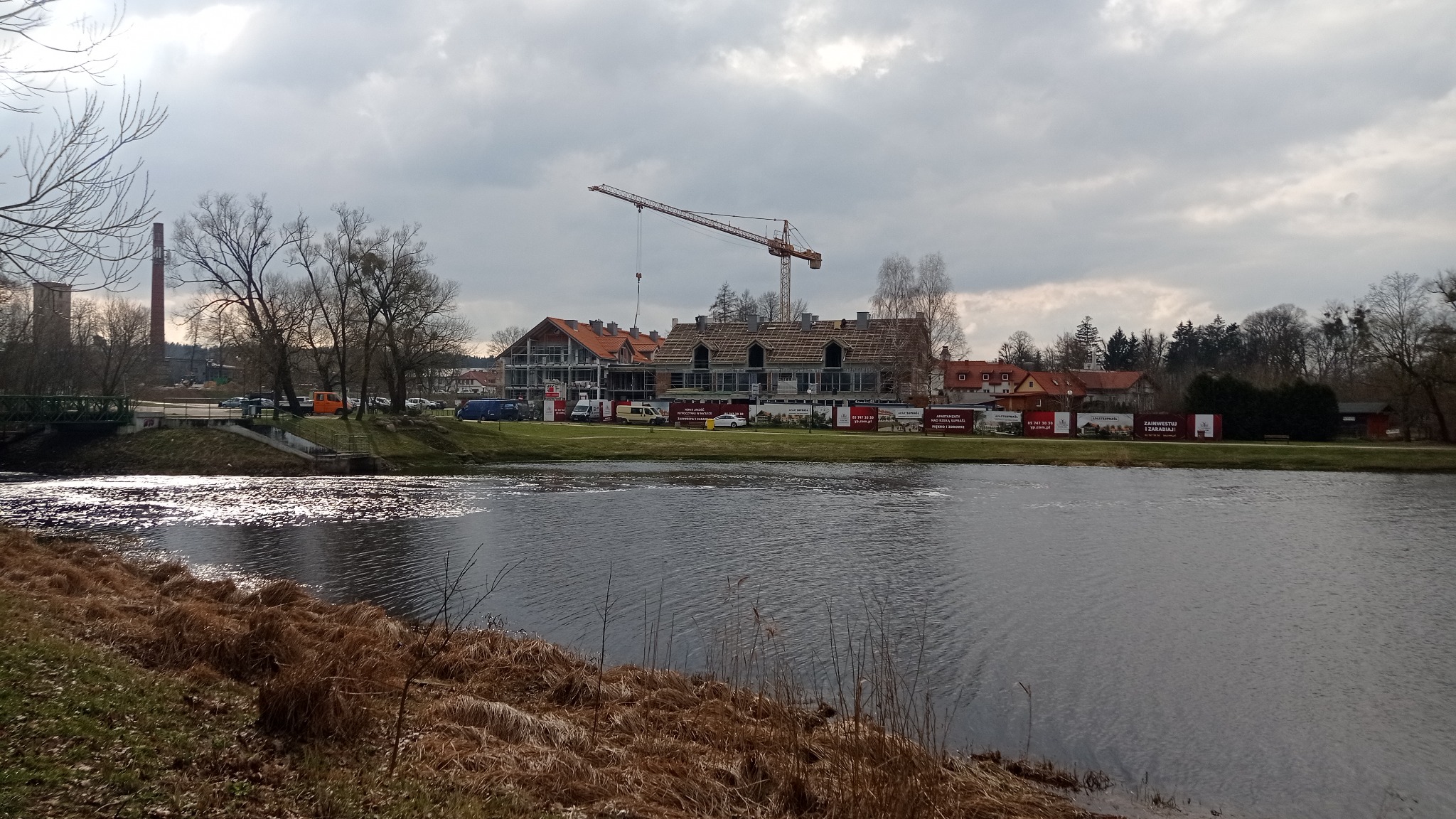 Budowa budynku hotelowego z usługami w Supraślu, widok zza rzeki, na pierwszym planie brzeg z suchą trawą, w tle budynek w stanie surowym otwartym z pomarańczową dachówką i żurawiem budowlanym.