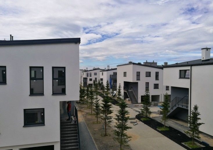 Osiedle domów jednorodzinnych w zabudowie szeregowej z białymi elewacjami i czarnymi ramami okiennymi, widoczne schody zewnętrzne i młode drzewka iglaste wzdłuż ścieżki.