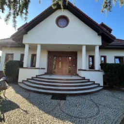 Elewacja domu jednorodzinnego z kolumnami, okrągłym oknem i schodami wyłożonymi kamieniem, widok z poziomu chodnika.