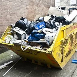 Żółty, mocno zabrudzony kontener na śmieci, wypełniony po brzegi różnymi odpadami, w tym workami na śmieci, elementami wyposażenia wnętrz i elektroniką, ustawiony na chodniku przy ceglanym budynku.