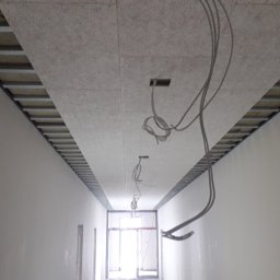 Adam - Montaż sufitu podwieszanego z widoczną konstrukcją nośną i wystającymi przewodami elektrycznymi w korytarzu z pomalowanymi na jasny kolor ścianami.