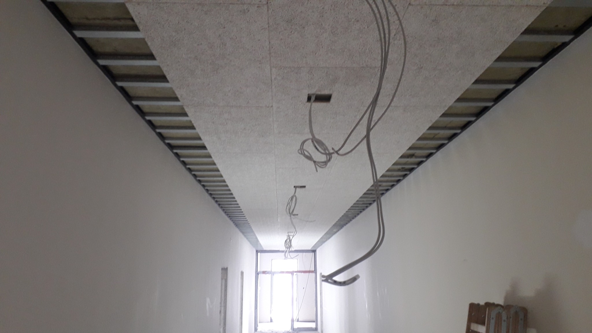 Montaż sufitu podwieszanego z widocznymi elementami konstrukcyjnymi i przewodami elektrycznymi w długim korytarzu, w trakcie prac wykończeniowych.