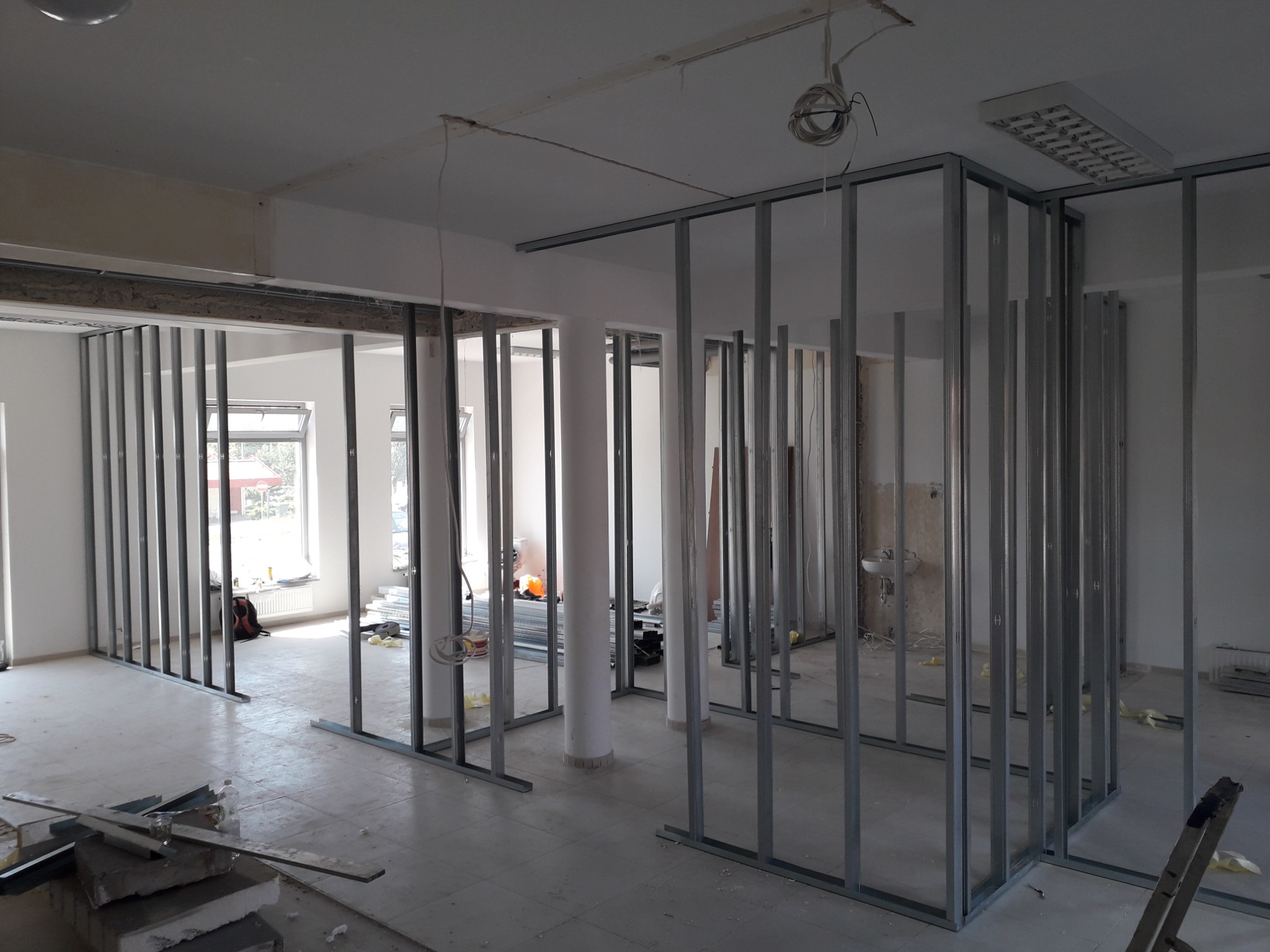 Wnętrze biura w trakcie remontu, widoczne metalowe profile ścian działowych, białe kolumny i surowy stan wykończenia podłogi.