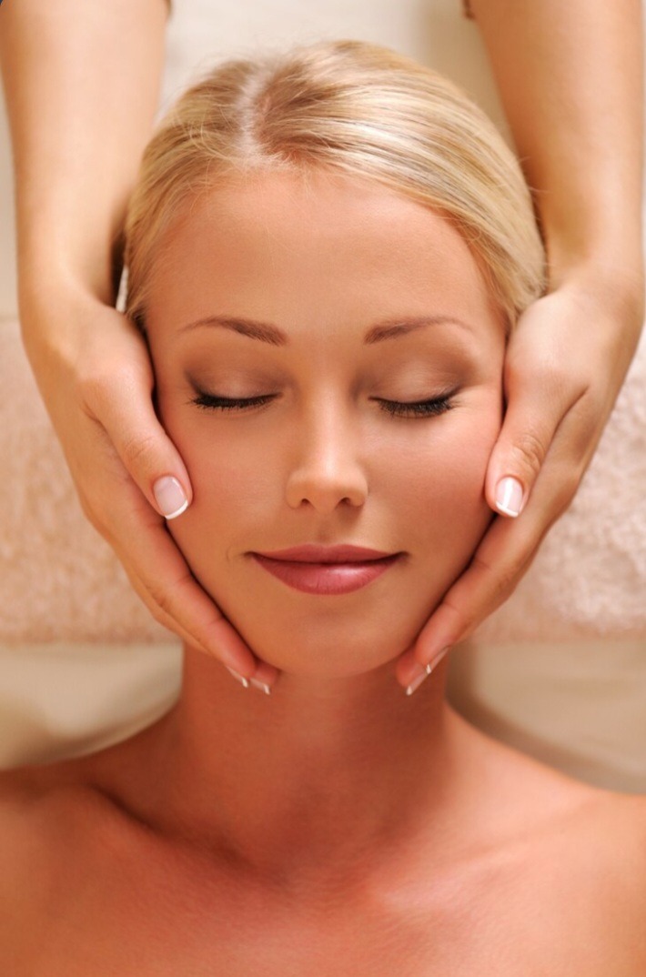 Kobieta z zamkniętymi oczami relaksuje się podczas masażu twarzy wykonywanego przez masażystę, widoczne dłonie masażysty delikatnie dotykają jej policzków i szyi.