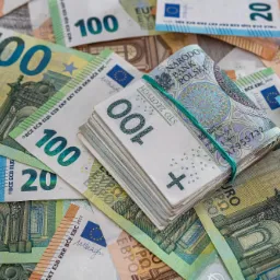 Stos banknotów polskich złotych spięty gumką na tle rozrzuconych banknotów euro o różnych nominałach.