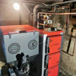Nowoczesny piec na paliwo stałe marki PAREX z podajnikiem, z widocznymi certyfikatami Eco Design i Klasa 5, zainstalowany w piwnicy z widoczną instalacją hydrauliczną.