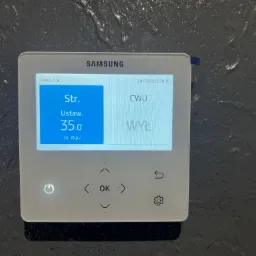 Biały panel sterowania Samsung z ekranem dotykowym, wyświetlający ustawienia temperatury ogrzewania na tle szarej, teksturowanej ściany.