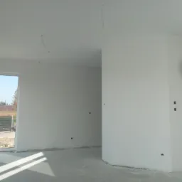 Surowe wnętrze nowo wykończonego pokoju z białymi ścianami, widoczne wystające kable z sufitu, okno z widokiem na zewnątrz i betonowa podłoga w szarym odcieniu.