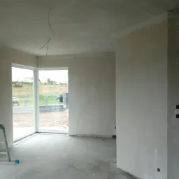 Wnętrze w Lubięcinie w trakcie prac przygotowawczych: surowe ściany po szpachlowaniu, widoczne kable wystające z sufitu, surowy beton na podłodze, drabina oparta o ścianę przy oknie.