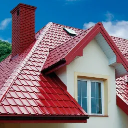 Czerwony dach z blachy trapezowej na domu z oknem mansardowym i ceglanym kominem, widok z dołu na błękitne niebo.