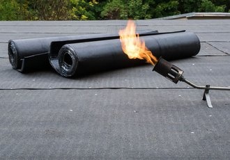 Prace dekarskie: palnik dekarski podgrzewa papę na płaskim dachu. Widoczne dwie rolki czarnej papy bitumicznej.