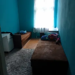 Pokój przed remontem: Dwa tapczany, turkusowe ściany, szary dywanik i okno z białymi firankami.