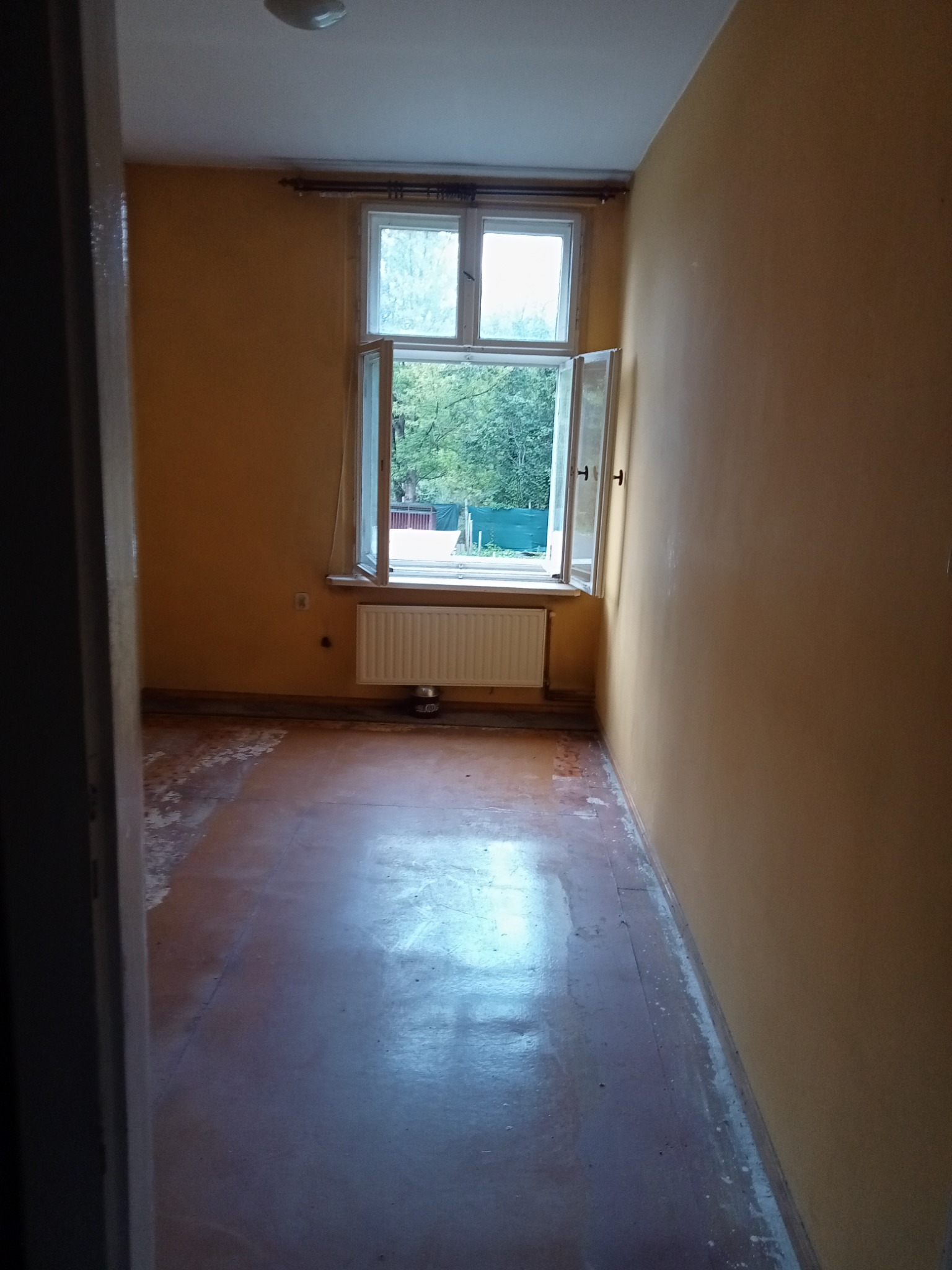 Wnętrze pokoju z otwartym oknem, widoczne ślady po usuniętej ścianie lub tapetach, odsłonięta podłoga przed remontem.