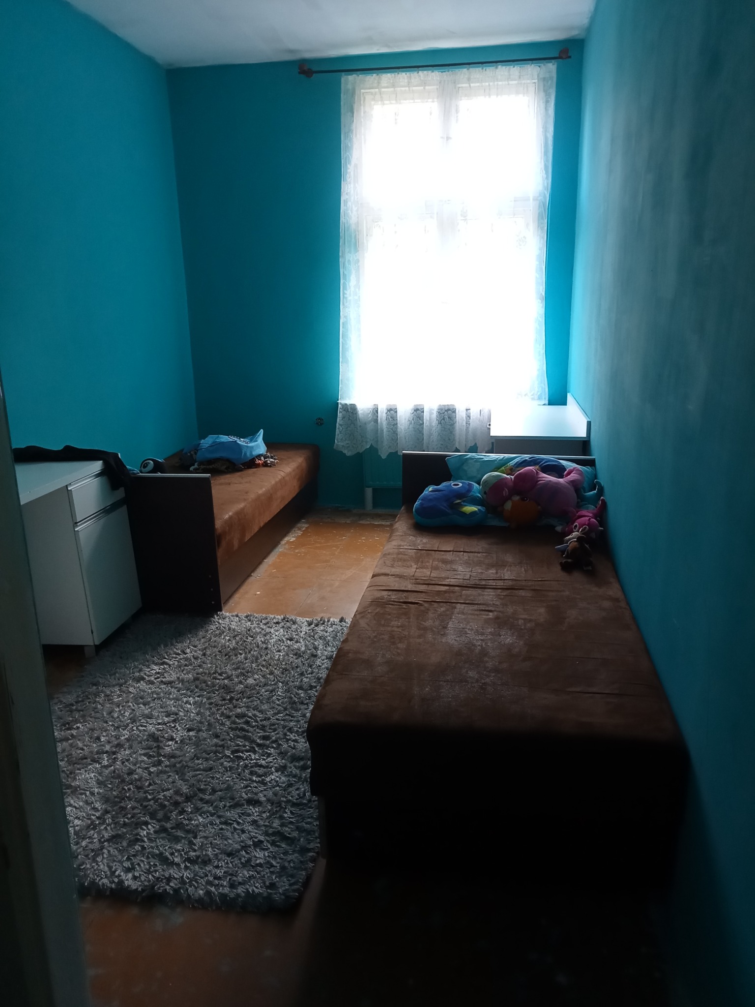 Pokój przed remontem: Dwa tapczany, turkusowe ściany, szary dywanik i okno z białymi firankami.