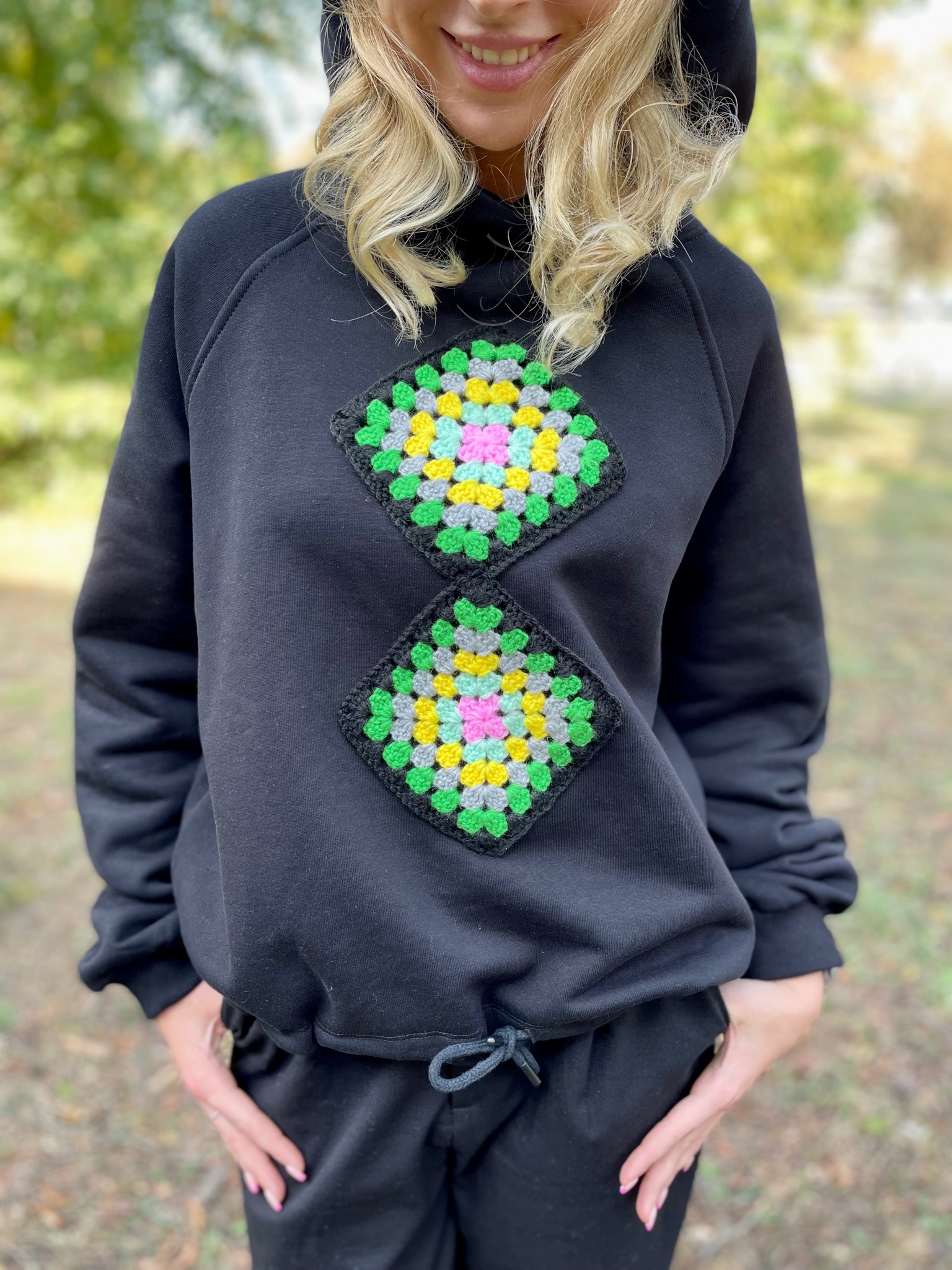 Czarna bluza z kapturem ozdobiona dwoma kwadratowymi, kolorowymi aplikacjami szydełkowymi w stylu granny square, noszona przez uśmiechniętą blondynkę na tle parkowej zieleni.