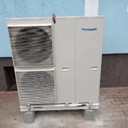 Zewnętrzna jednostka pompy ciepła Panasonic na betonowych podstawach przed budynkiem z niebieską elewacją i szarym cokołem.