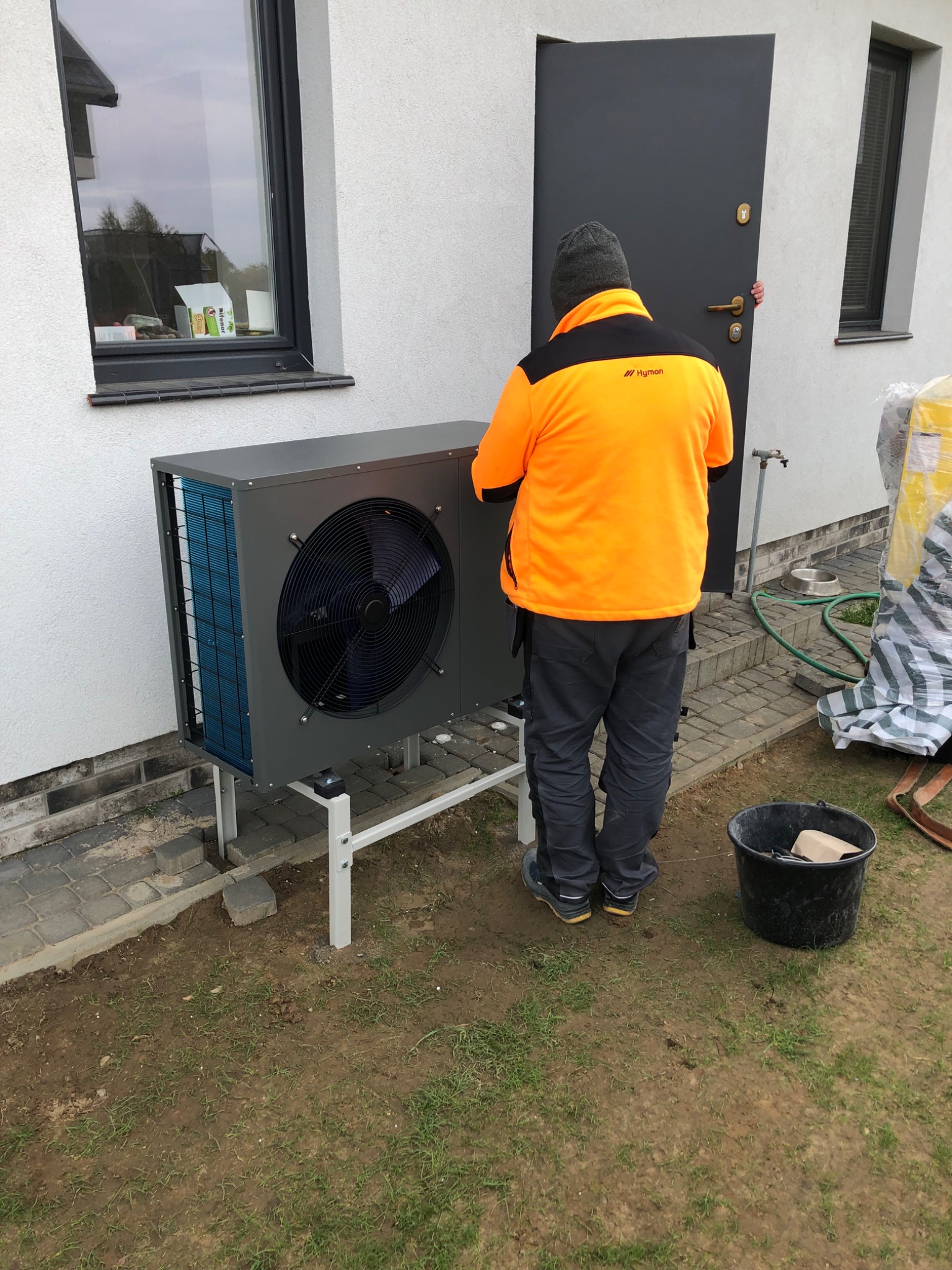 Szara pompa ciepła typu monoblok na białym stelażu, zamontowana przy elewacji domu. Widoczny pracownik w pomarańczowej kurtce z logo firmy, oraz kubeł z narzędziami obok.