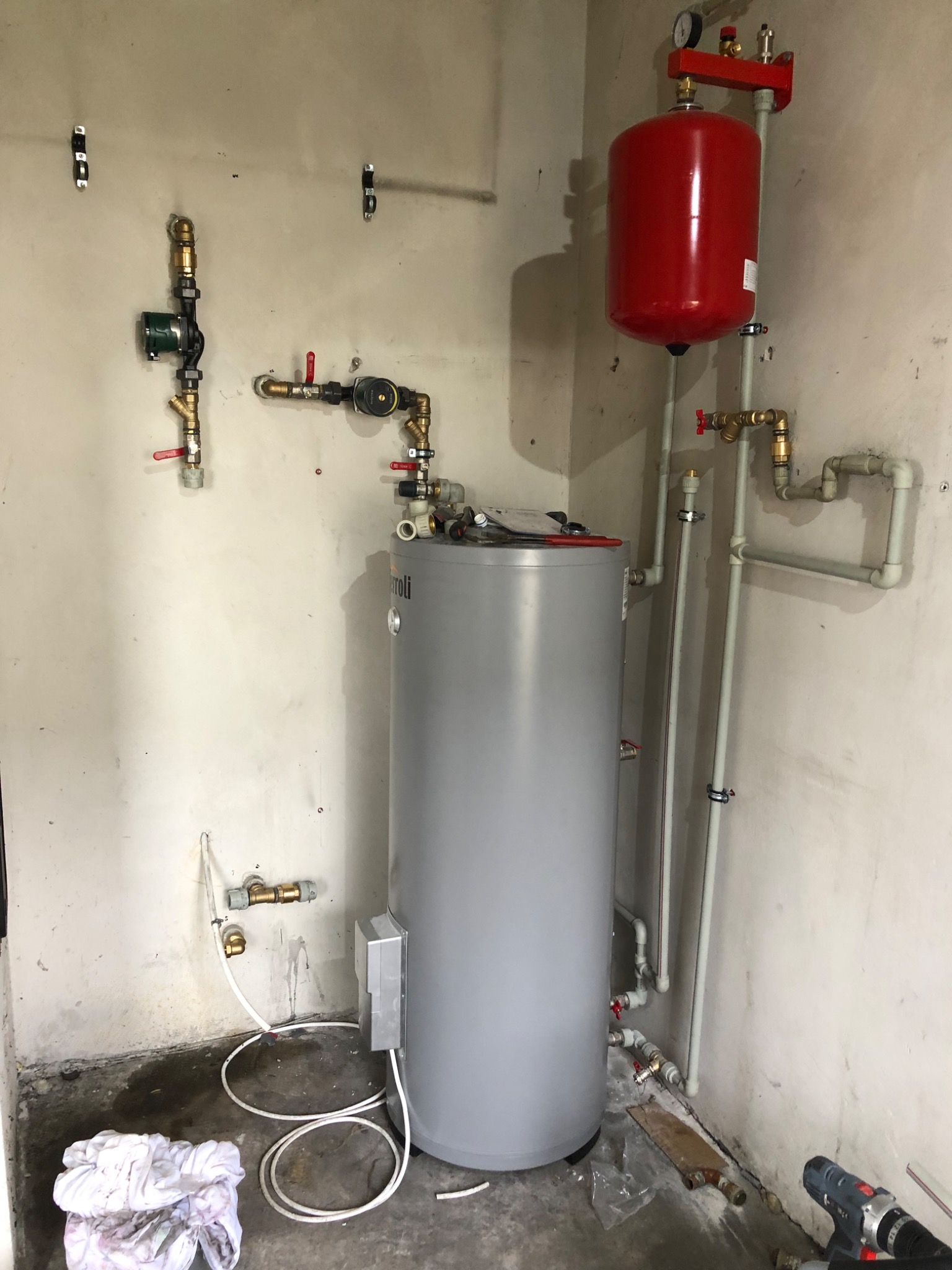 Szary, pionowy zasobnik ciepłej wody użytkowej z podłączonymi rurami miedzianymi i plastikowymi, czerwony zbiornik wyrównawczy na ścianie, widoczne narzędzia i ślady po instalacji na surowej ścianie.