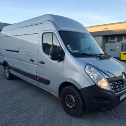 Srebrny bus Renault Master z włączonymi światłami, zaparkowany na szarym asfalcie przed budynkiem. Na desce rozdzielczej widoczny czarny panel z dodatkowymi światłami.