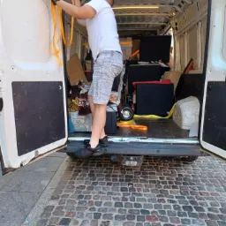 Mężczyzna w czarnych rękawicach zabezpiecza pomarańczowymi pasami ładunek wewnątrz białego busa, widoczne kartony, meble i inne przedmioty.