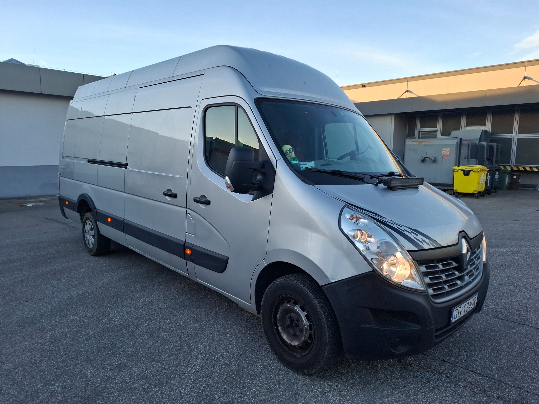 Srebrny bus Renault Master z włączonymi światłami, zaparkowany na szarym asfalcie przed budynkiem. Na desce rozdzielczej widoczny czarny panel z dodatkowymi światłami.