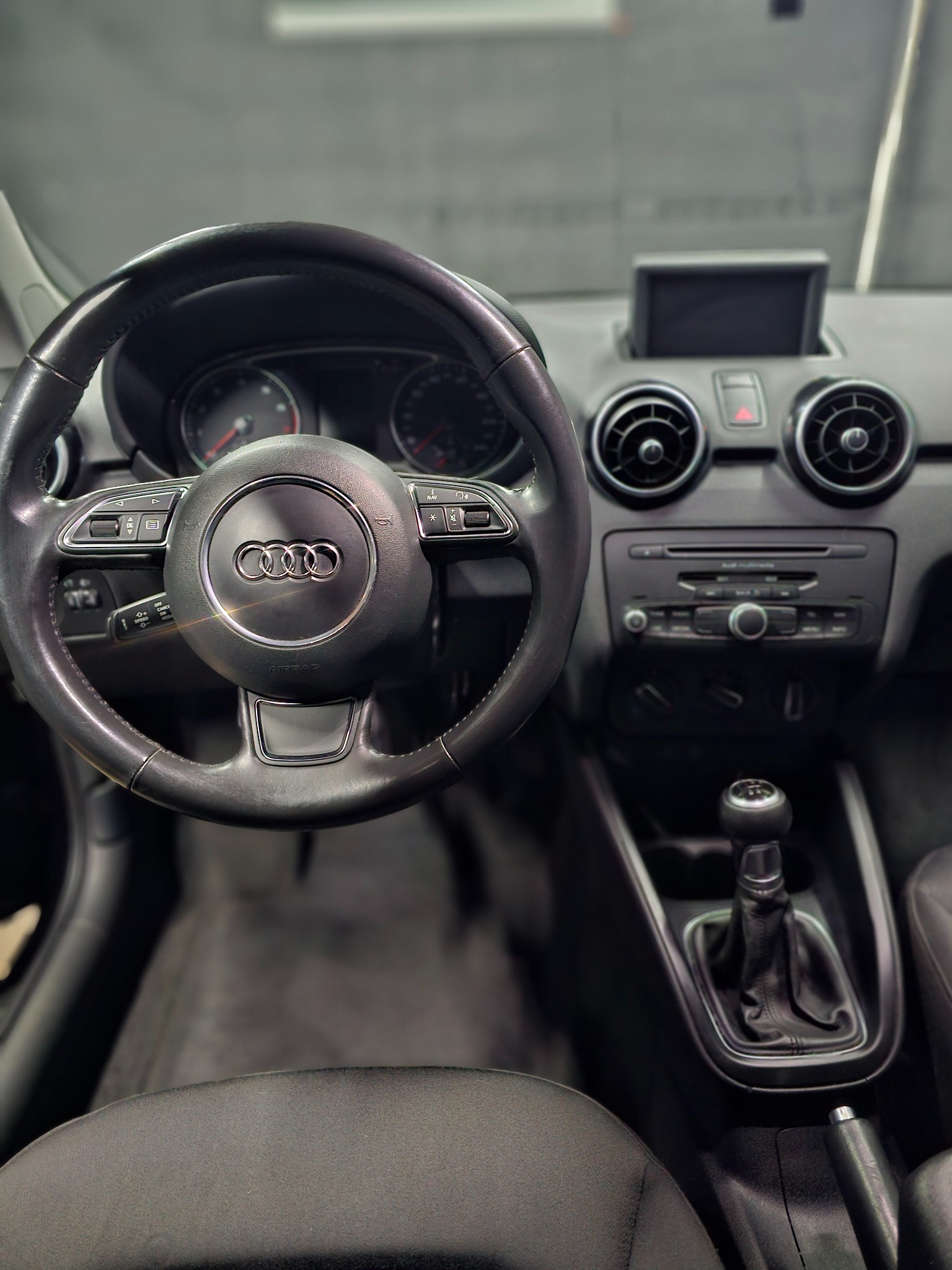 Wnętrze samochodu Audi A1 z widoczną kierownicą z logo, deską rozdzielczą, radiem i dźwignią zmiany biegów, z perspektywy kierowcy.