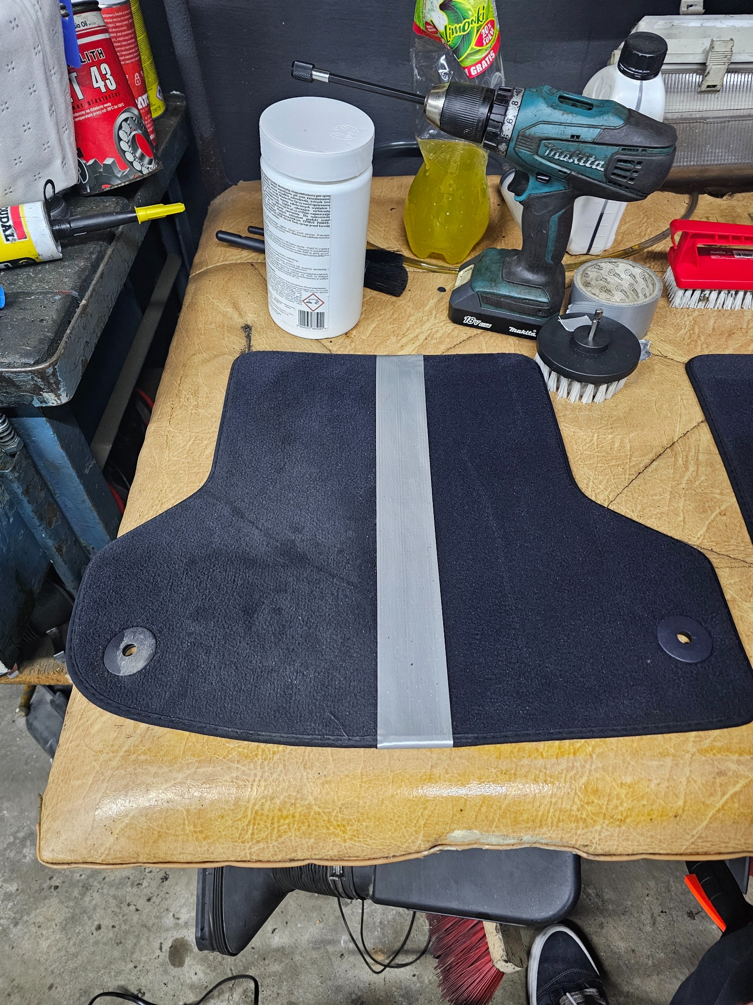 Czyszczenie dywanika samochodowego z użyciem wkrętarki z szczotką, widoczny środek czyszczący i inne akcesoria na warsztatowym stole.