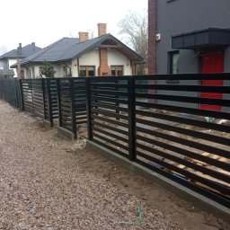 JarBud - Nowoczesne ogrodzenie panelowe z poziomych, czarnych listew, zamontowane na betonowej podmurówce, widok z zewnątrz posesji, z kamienistym podłożem i domami w tle.