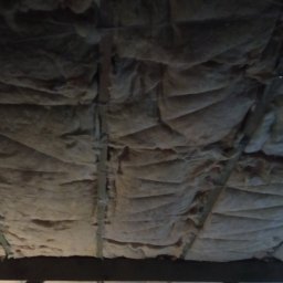 JarBud - Niedostatecznie oświetlone poddasze z widoczną wełną mineralną pomiędzy drewnianymi belkami stropowymi, zabezpieczoną sznurkiem.