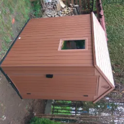 Brązowy domek narzędziowy z tworzywa sztucznego, ustawiony na ziemi obok ogrodzenia i drzew, z widocznym stosem drewna i elementami ogrodowymi w tle.