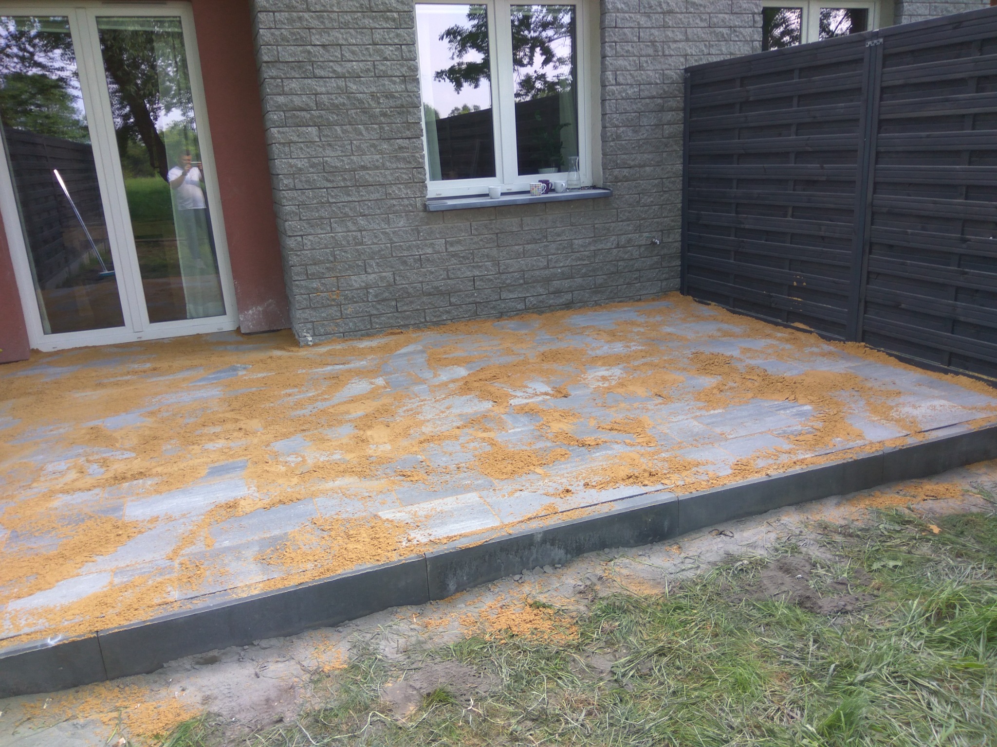 Szare płyty tarasowe z obrzeżami, posypane piaskiem, widok na szary mur z oknem i ciemne, poziome ogrodzenie.