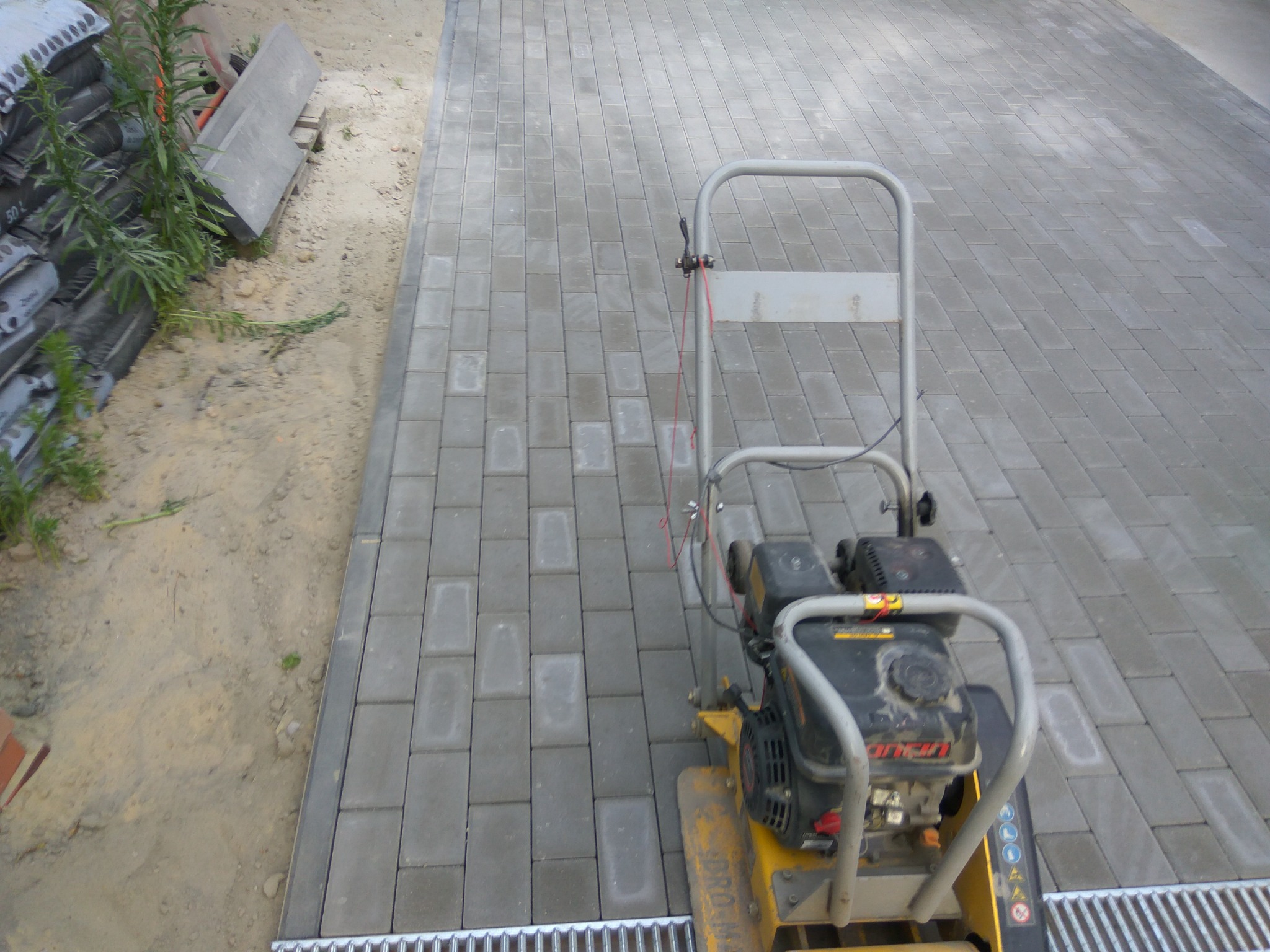 Wibro ubijak na świeżo ułożonej szarej kostce brukowej, widoczne worki z materiałem i elementy odwodnienia liniowego.