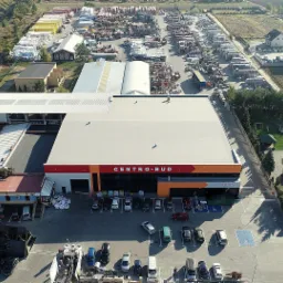 Widok z drona na duży skład budowlany Centro-Bud z rozległym placem składowym pełnym materiałów budowlanych, parkingiem z samochodami i otaczającą zabudową jednorodzinną.