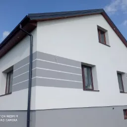 Dwukondygnacyjny dom z białymi ścianami, szarym pasem z białymi paskami i szarym cokołem, brązowe ramy okienne, widok z zewnątrz, błękitne niebo w tle.