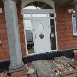 PHU SEBMARK Marek Olejnik - Białe drzwi wejściowe z owalnym przeszkleniem, zamontowane w surowym stanie budynku z cegły, widoczne kolumny i okna, w tle zaparkowany samochód dostawczy.