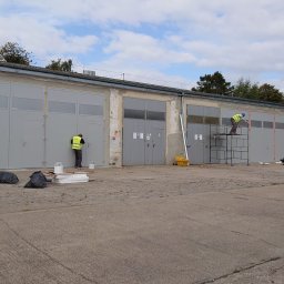 PHU SEBMARK Marek Olejnik - Malowanie szarych drzwi garażowych: Dwóch pracowników w kamizelkach odblaskowych maluje metalowe drzwi garażowe na zewnątrz budynku, jeden z drabiny, drugi z ziemi. Widoczne worki na śmieci, puszki...