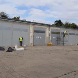 Malowanie szarych drzwi garażowych: Dw&oacute;ch pracownik&oacute;w w kamizelkach odblaskowych maluje metalowe drzwi garażowe na zewnątrz budynku, jeden z drabiny, drugi z ziemi. Widoczne worki na śmieci, puszki...