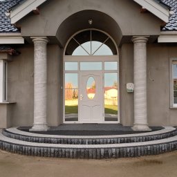 PHU SEBMARK Marek Olejnik - Białe drzwi wejściowe z owalnym przeszkleniem, otoczone kolumnami i łukiem, widoczne z zewnątrz na tle domu z szarą elewacją i schodami z kostki brukowej.