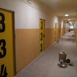 PHU SEBMARK Marek Olejnik - Długi korytarz z żółtymi drzwiami oznaczonymi czarnymi numerami, widoczne ślady remontu, kartony z narzędziami na podłodze, malowanie ścian w dwóch kolorach: biały i żółty.
